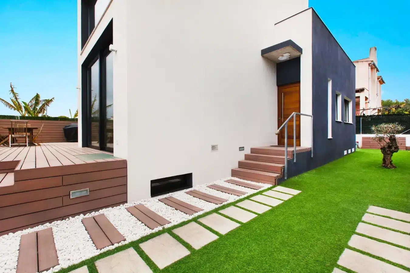 Tarima exterior marrón con césped artificial y pasos de piedra blanca en vivienda moderna
